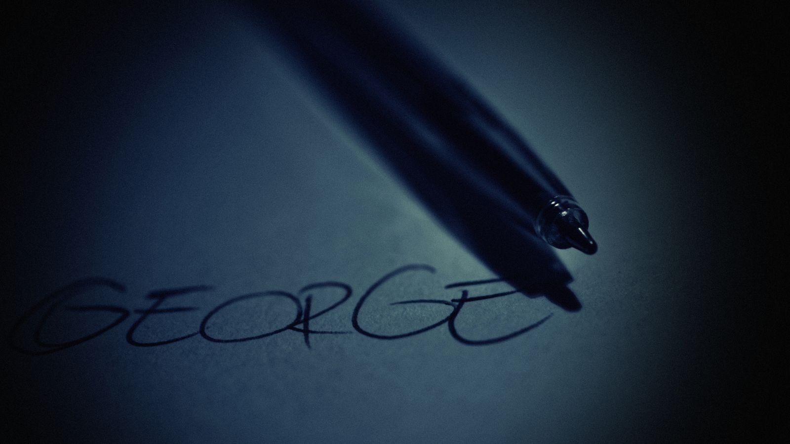 Artistic close-up of a pen casting shadow on handwritten name 'George' in soft dark lighting.