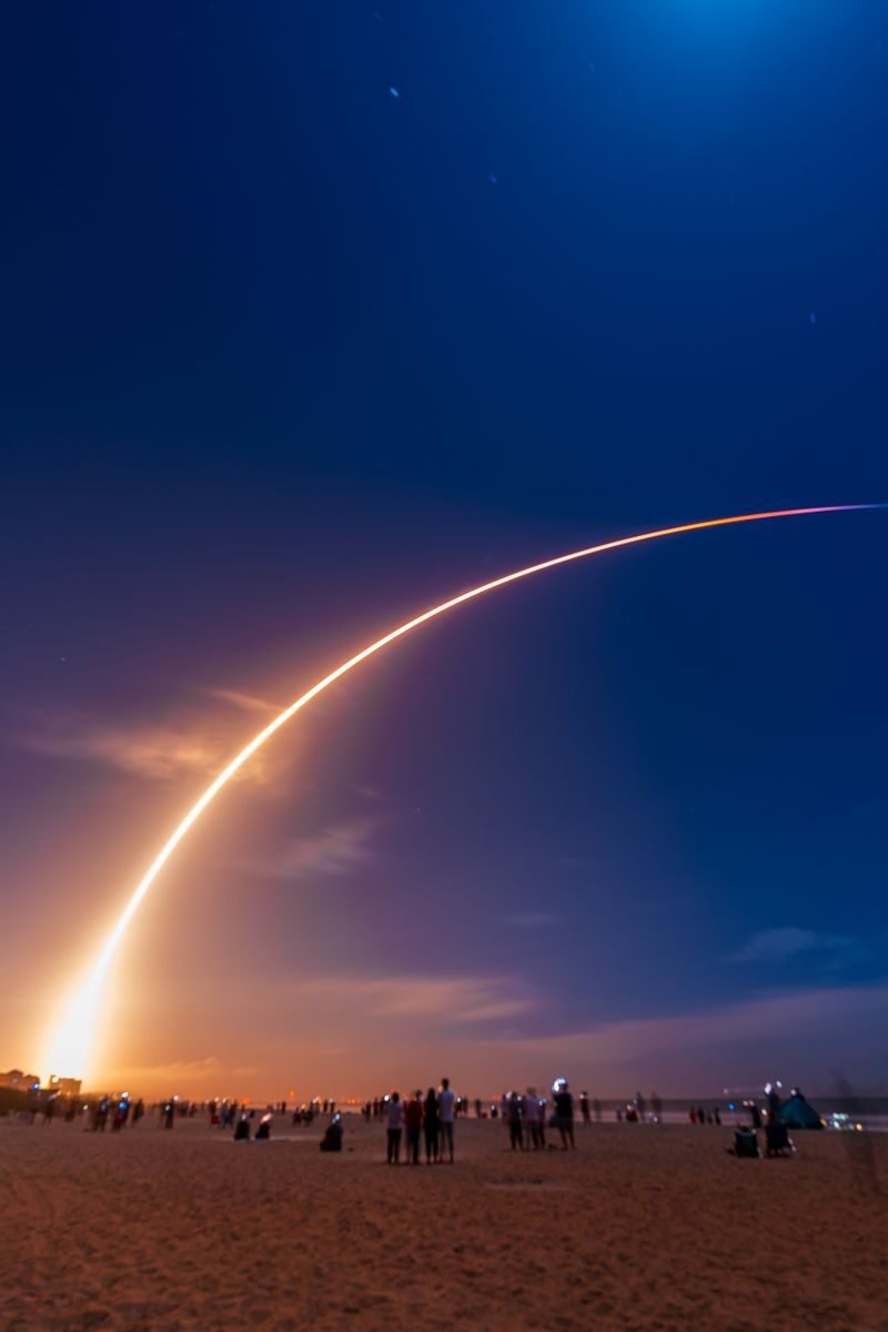 A stunning rocket launch leaving a bright arc above a beach with onlookers.