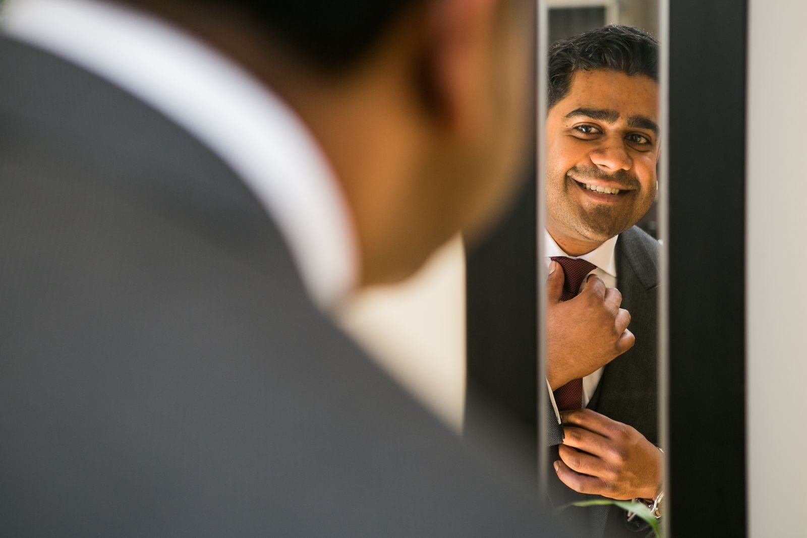 Crop ethnic male in elegant suit preparing for special festive event while adjusting tie and looking at mirror