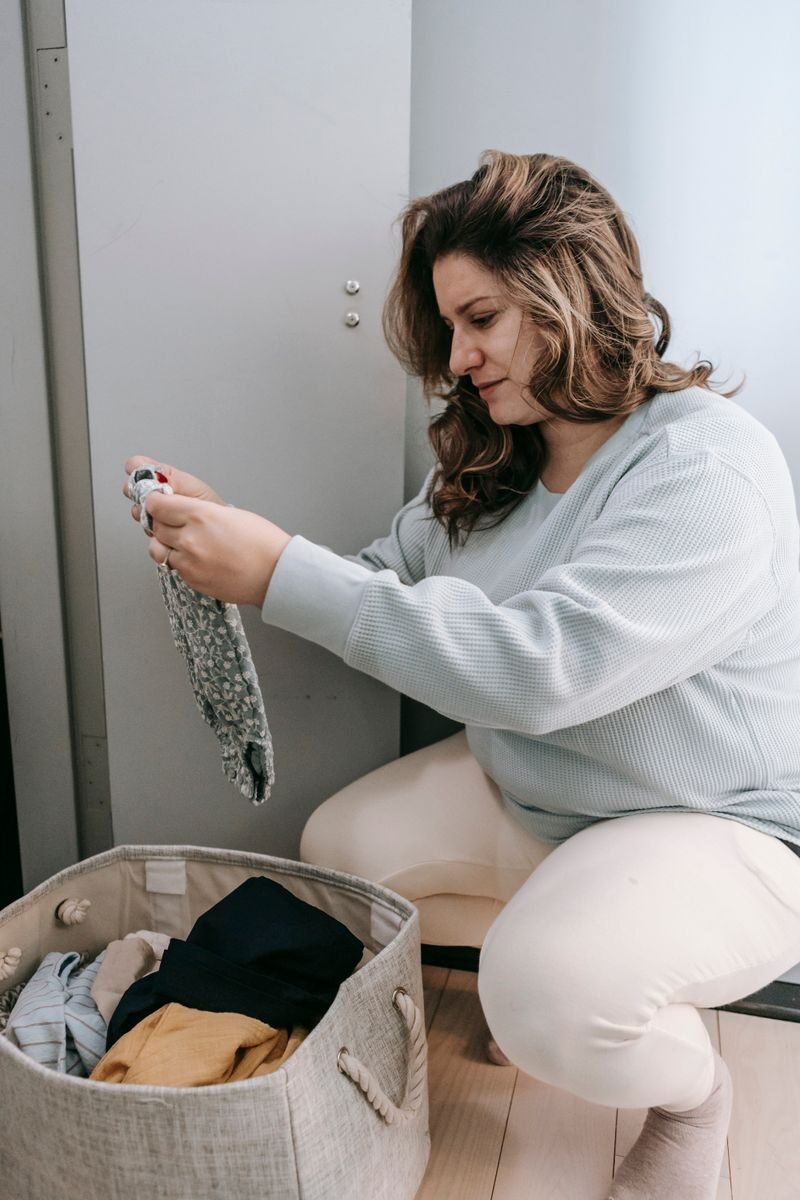 Focused female in casual wear picking dirty clothes from laundry basket while sitting in light room at home during daily housework routine