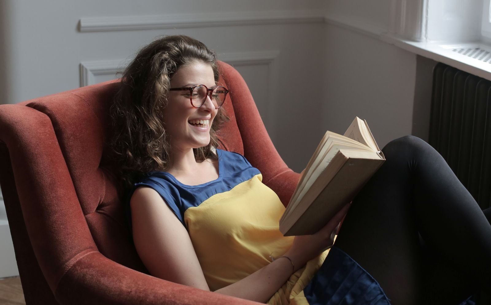 Positive young lady in casual wear and eyeglasses sitting with crossed legs on cozy chair while reading book and laughing in apartment