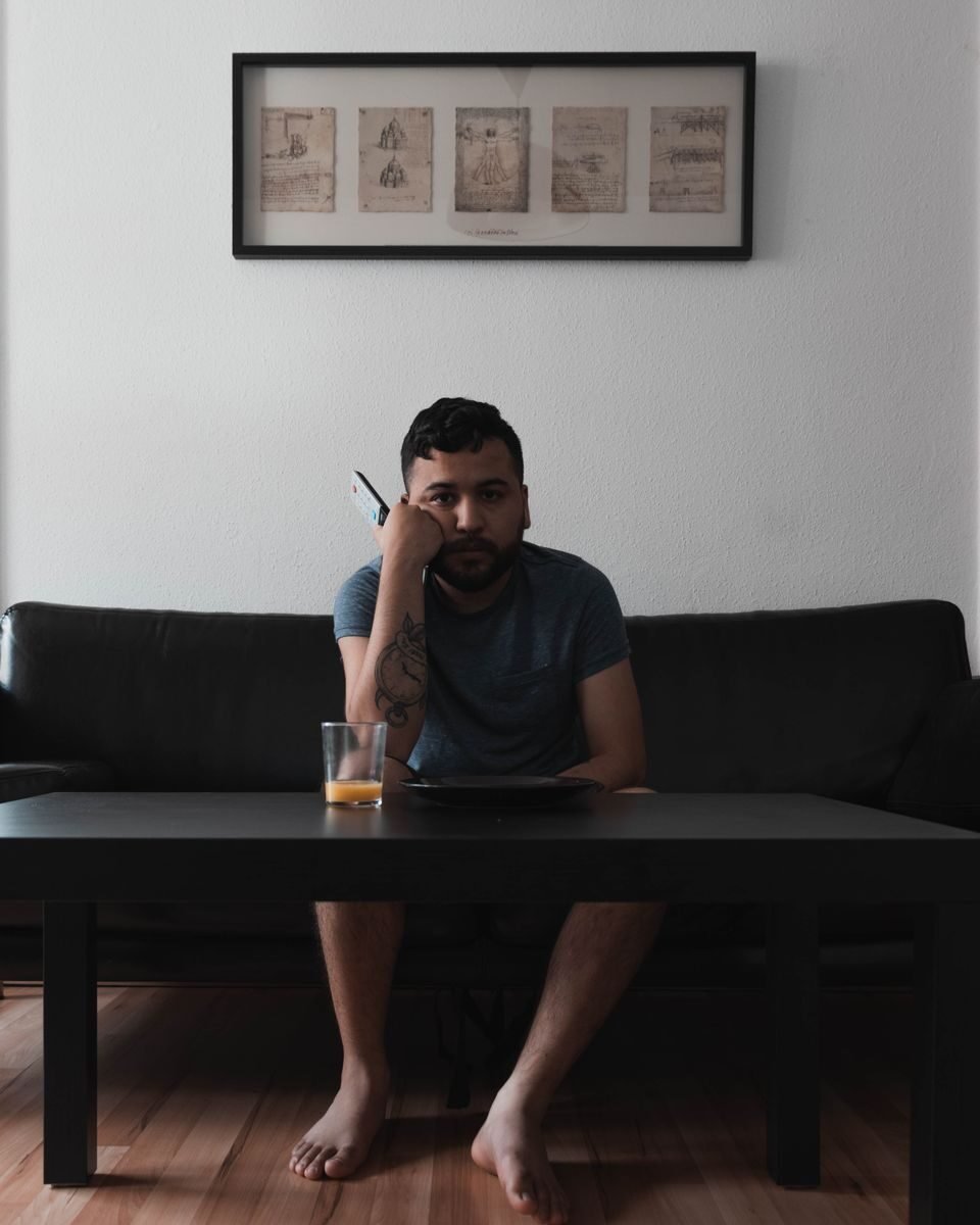 An adult man sitting on a sofa indoors holding a remote, juice on table.