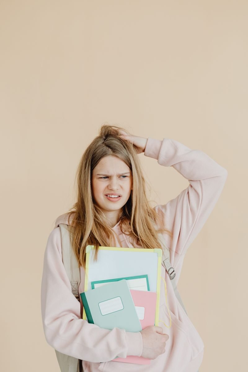 Teenage girl on beige backdrop holding colorful notebooks, contemplating something.