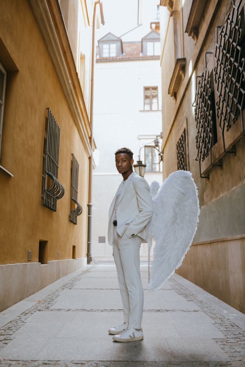 A man in a white suit with angel wings standing in a narrow urban alleyway, exuding divine elegance.