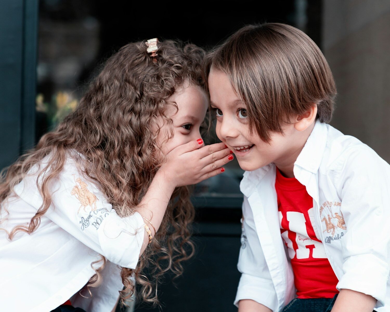 A girl with long curly hair whispering into the ear of a boy
