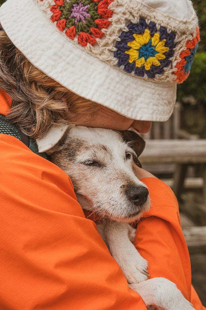 Person in orange jacket hugs a dog wearing a hat